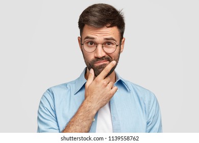 Image of hesitant unshaven European male with thick beard, holds chin, purses lips with clueless expressions, doubts what to eat delicious for supper, isolated over white background. What to choose?