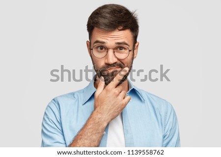 Image of hesitant unshaven European male with thick beard, holds chin, purses lips with clueless expressions, doubts what to eat delicious for supper, isolated over white background. What to choose? Zdjęcia stock © 