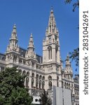 The image highlights the dramatic central tower (Mittelturm), crowned by the famous Rathausmann statue, soaring into a crisp, clear blue sky. Elaborate stone tracery, pointed arches, fine sculptures, and intricate spires showcase the exceptional craftsmanship of 19th-century European builders. Lush green foliage frames the lower left side, creating a natural contrast against the pale limestone structure, while vibrant red flowers decorate the balcony ledges.