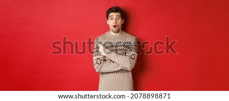 Image of impressed handsome guy, showing awesome christmas promo offer, pointing finger at upper left corner and looking amazed, standing in xmas sweater over red background