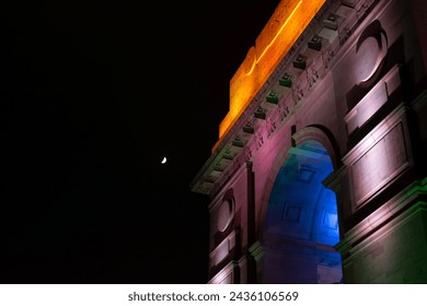 Image of India Gate at night with colors projected on it and moon in background