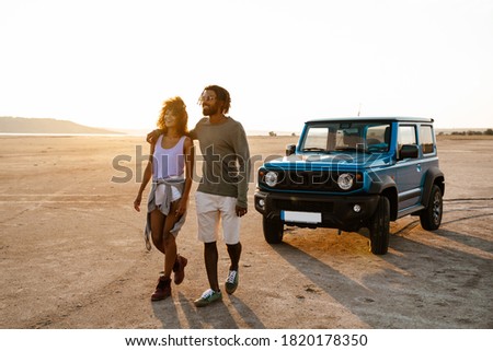 Image of joyful african american couple hugging and walking while travelling with car on desert