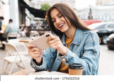 Image Of Joyful Brunette Woman Playing Video Game On Mobile Phone While Sitting At Street Cafe Outdoors