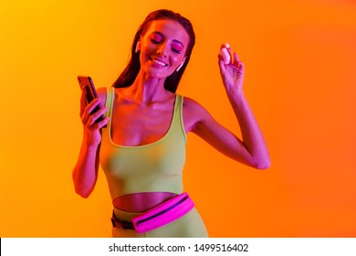 Image Of Joyful Brunette Woman Wearing Formfitting Sportswear Dancing While Using Smartphone And Earpods Isolated Over Orange Neon Wall