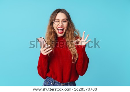 Image of joyful caucasian woman holding cellphone and gesturing ok sign while winking isolated over blue background