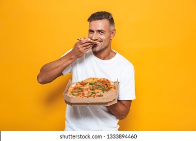 Image of joyful man 30s in white t-shirt holding and eating pizza while standing isolated over yellow background