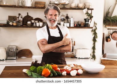 Image Of Joyful Mature Man In Apron Winking And Showing Thumb Up While Standing At Cozy Kitchen