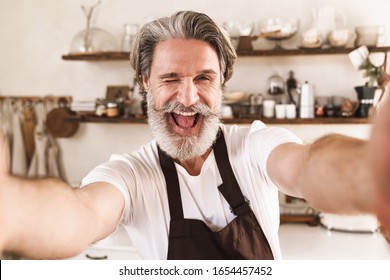 Image Of Joyful Mature Man In Apron Winking And Smiling While Taking Selfie At Cozy Kitchen