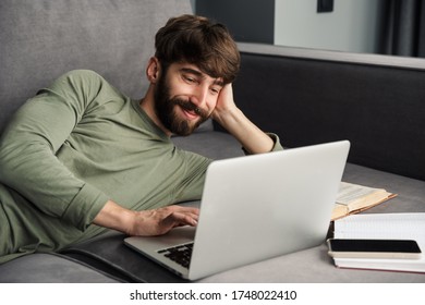 Image of joyful young man in casual wear using laptop and smiling while lying on sofa in living room - Shutterstock ID 1748022410