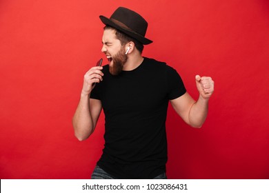 Image of joyous guy wearing black t-shirt and hat singing and listening to music with cell phone and wireless earphones isolated over red wall