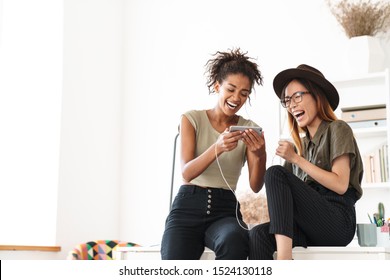 Image Of A Laughing Emotional Happy Positive Two Women Colleagues In Office Using Mobile Phone Listening Music With Earphones.