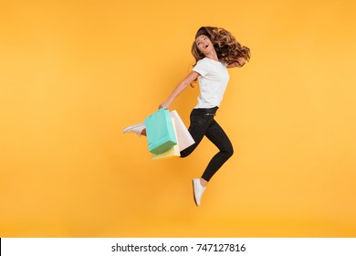 Image of laughing pretty young woman jumping isolated over yellow wall background. Looking aside holding shopping bags.