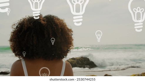 Image of light bulb icons over african american woman by seaside. Global social media, networks, digital interface and data processing concept digitally generated image. - Powered by Shutterstock