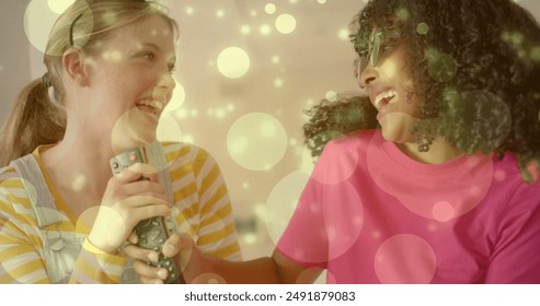 Image of light spots over two happy diverse teenage girls miming singing with tv remote control. Friendship, youth, free time, fun, music, weekend and lifestyle, digitally generated image. - Powered by Shutterstock