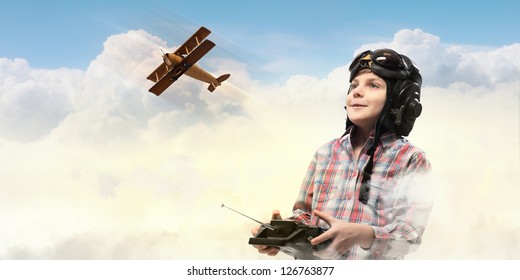 Image of little boy in pilots helmet playing with toy radiocontrolled airplane against clouds background