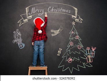 Image of little child wearing hat standing on stool near Christmas tree while drawing on blackboard. - Powered by Shutterstock