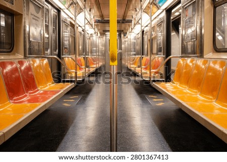 An image looking down the length of a New York subway train. Orange and red sets line either side of the carriage with metal poles standing in the centre.