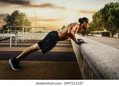 Image of a man doing incline push-ups in an outdoor training park during sunset. He is wearing black shorts and training gloves. The background shows exercise equipment and a slight mist.  - Powered by Shutterstock