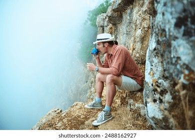 Image of a man with a hat drinking from a bottle, sitting on a rocky cliff edge in the highlands, surrounded by mist and rugged terrain. Adventure and solitude. - Powered by Shutterstock