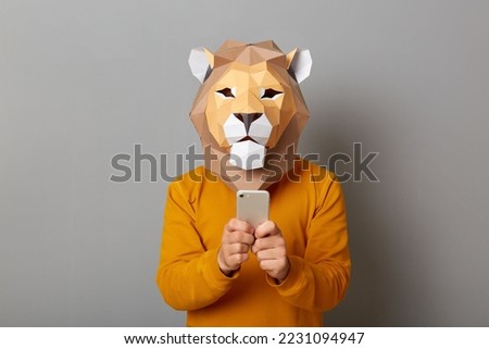 Image of man with lion mask with wearing orange sweater, standing with cell phone in hands, using mobile phone for browsing Internet or chatting in social networks posing isolated over gray background
