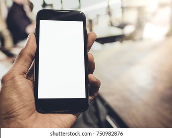 Image Of Man's Hand Is Holding A Black Cell Phone With Blank White Screen And Blur Silver Laptop On Wood Table In Coffee Cafe  Background