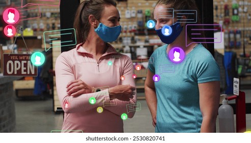 Image of media icons over two caucasian female workers with face masks talking. social media and communication interface during covid 19 pandemic concept digitally generated image. - Powered by Shutterstock