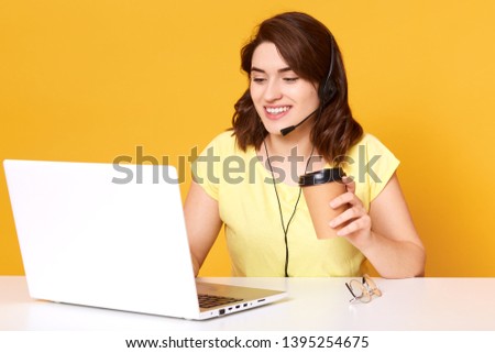 Image of miling freelance operator working online with headsets and laptop computer at office or home. Cheerful call center female talking with customer and drinking coffee, beind in good mood.