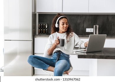 밝은 부엌에 앉아 있는 동안 노트북을 사용하여 헤드폰을 끼고 있는 현대 흑인 소녀의 이미지 스톡 사진