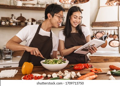 Image of multicultural couple smiling and reading book with recipes while cooking dinner in cozy kitchen - Powered by Shutterstock