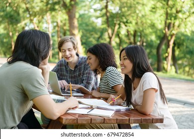 Image d'un groupe multiethnique de jeunes étudiants joyeux assis et étudiant à l'extérieur en utilisant un ordinateur portable. En regardant de côté. : photo de stock
