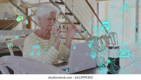 Image of network of connections with icons over senior caucasian woman using laptop. global connections, digital interface and technology concept digitally generated image.