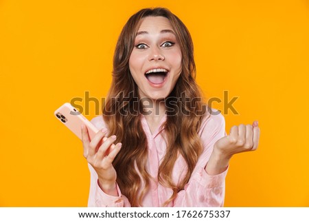 Image of nice excited woman making winner gesture and using mobile phone isolated over yellow background
