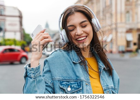 Image of nice laughing woman listening music with smartphone and wireless headphones on city street