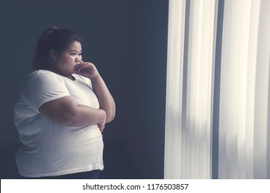 Image of obese woman looks pensive while standing by the window 