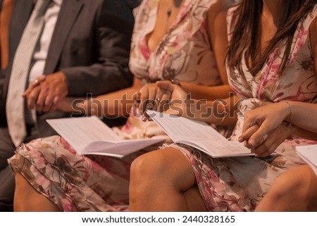 The image offers a close-up of elegantly dressed attendees seated with event programs in hand. The focus on the hands and attire suggests anticipation and engagement, characteristic of an audience at
