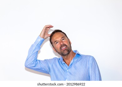 Image Of An Older Attractive Man In A Business Shirt Standing In Front Of A White Wall. He Is Smiling At The Camera And Scratching His Head. 