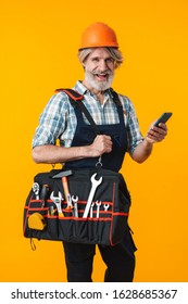 Image of optimistic happy elderly grey-haired bearded man builder in helmet posing isolated over yellow wall background holding bag with equipment instruments and mobile phone.