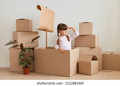 Image of optimistic little girl making ship with flag during relocation and playing sailor at new apartment, having fun while moving, posing surrounded packages with belongings.