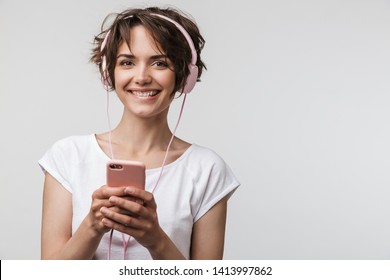 Image Of Optimistic Woman In Basic T-shirt Holding Smartphone While Listening To Music With Headphones Isolated Over White Background