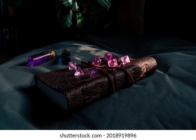 Image of pink transparent gaming dice on a leather-bound notebook partially lit by the sun. In the background two potions in glass stopper bottles