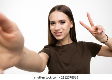 Image of pleased brunette woman laughing and showing peace sign while taking selfie photo on cellphone isolated over white background