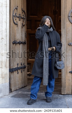 The image portrays a candid moment as a young woman, clad in a chic black coat and denim, steps out from the ancient doorway of a cathedral. With one hand delicately touching her face, she appears to