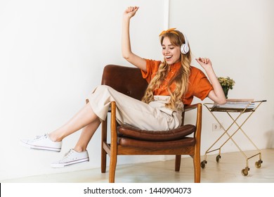 Image of a positive emotional smiling young blonde girl at home indoors listening music with headphonws sitting on chair dancing.