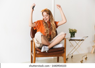 Image of a positive optimistic smiling young blonde girl at home indoors listening music with earphones sitting on chair dancing.