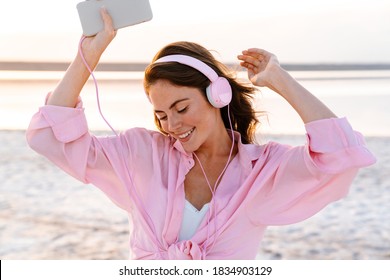 Image of a positive optimistic young girl in sunglasses listening music by headphones outdoors at the beach