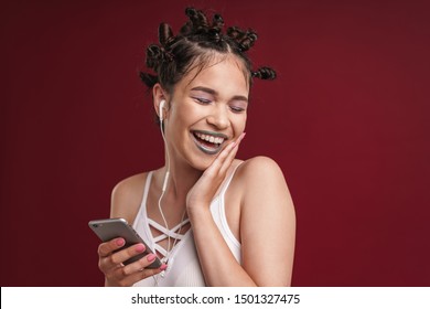 Image Of Positive Punk Girl With Bizarre Hairstyle And Dark Lipstick Laughing While Using Smartphone With Earphones Isolated Over Red Background