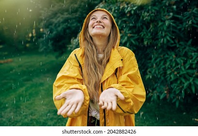 Image of a positive young blonde woman smiling wearing yellow raincoat during the rain in the park. Cheerful female enjoying the rain outdoors. Woman looking up and catching the rain drop with hands. - Powered by Shutterstock