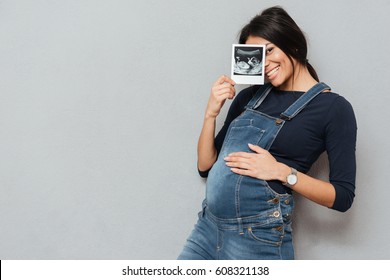 Image of pregnant smiling woman standing and posing while showing ultrasound scans over grey background. Looking at camera.
