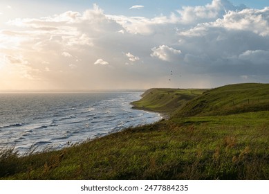 The image presents a coastal landscape during sunrise or sunset. The foreground features grassy terrain leading to a cliff edge, with the coastline extending into the distance. A few birds are visible - Powered by Shutterstock