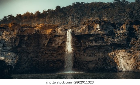 The image presents a dramatic, eye-level wide shot of a waterfall cascading over a rugged, vertical rock face. In the foreground, the dark, turbulent water of a pool receives the waterfall, with the surface reflecting the surrounding cliffs and sunlight. The middle ground is dominated by the massive cliff, which displays layered, earthy textures of ochre, brown, and grey stone, featuring sharp edges and crevices where the water flows in a concentrated, vertical stream. The sunlit upper ledge of the cliff provides a striking contrast, glowing with warm, golden hues against the deeper, shadowed crags below. The background rises above the cliff as a dense, dark forest of olive-green trees, set against a pale, bright sky that suggests a clear day. Natural, directional lighting illuminates the scene from the side, emphasizing the rugged geological textures and the dynamic motion of the white, splashing water as it hits the pool. The composition conveys a mood that is majestic, serene, and untamed, highlighting the raw power of the natural landscape.